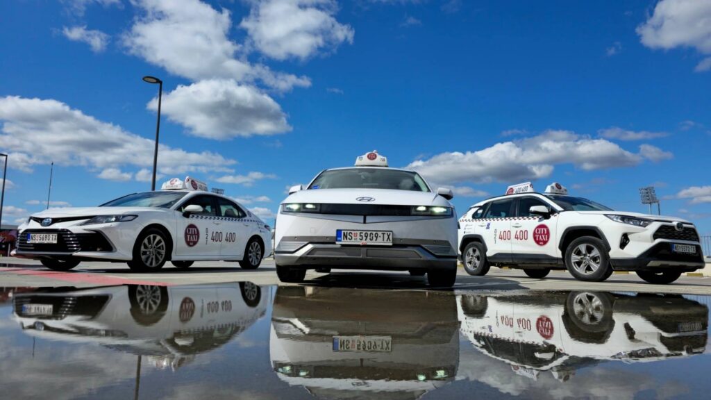 Tri moderna taxi vozila sa oznakom NS TAXI, parkirana na reflektujućoj površini, uz naglasak na ekološki pristup